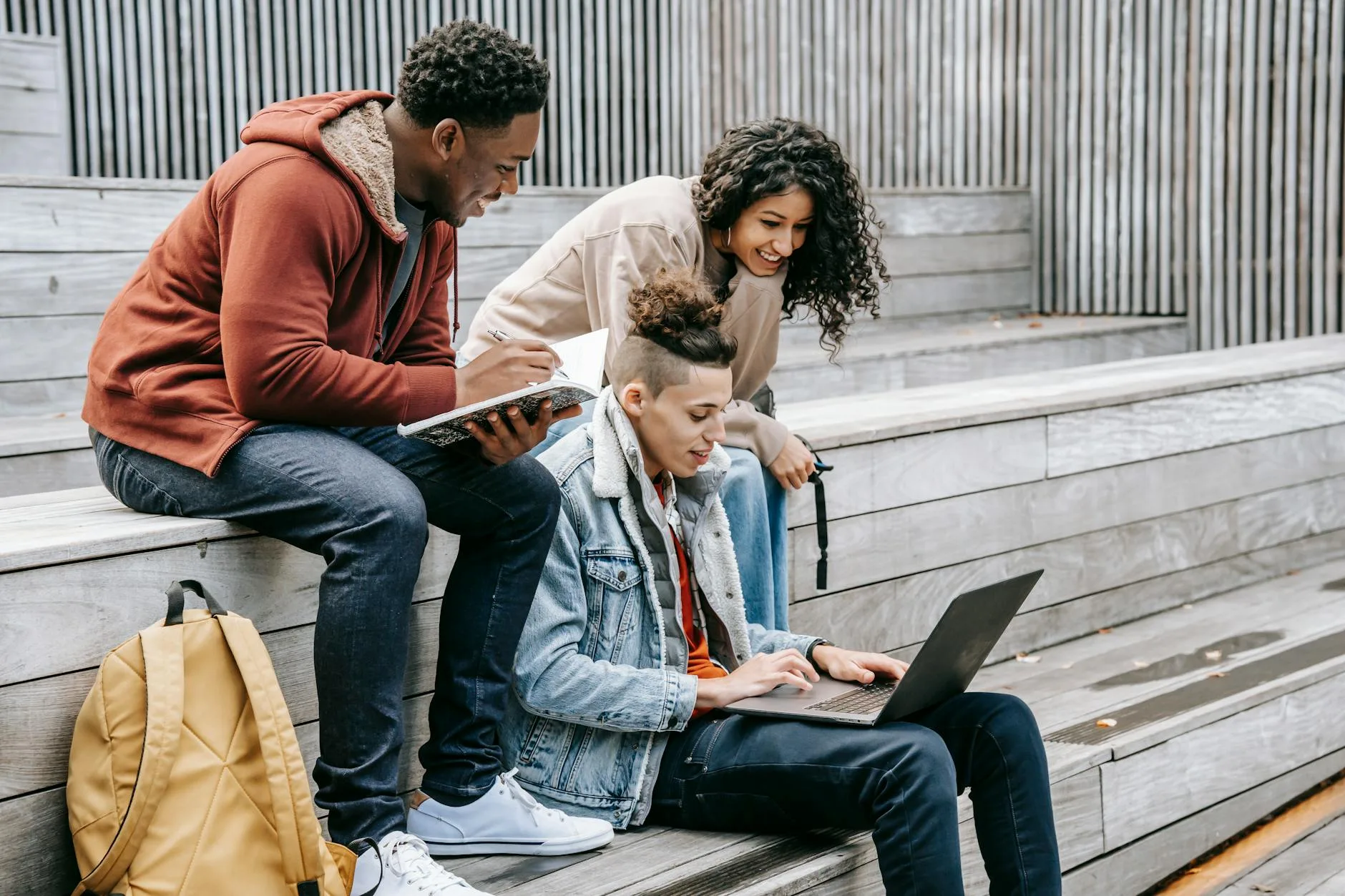 cheerful diverse students sharing laptop while studying on street stairs