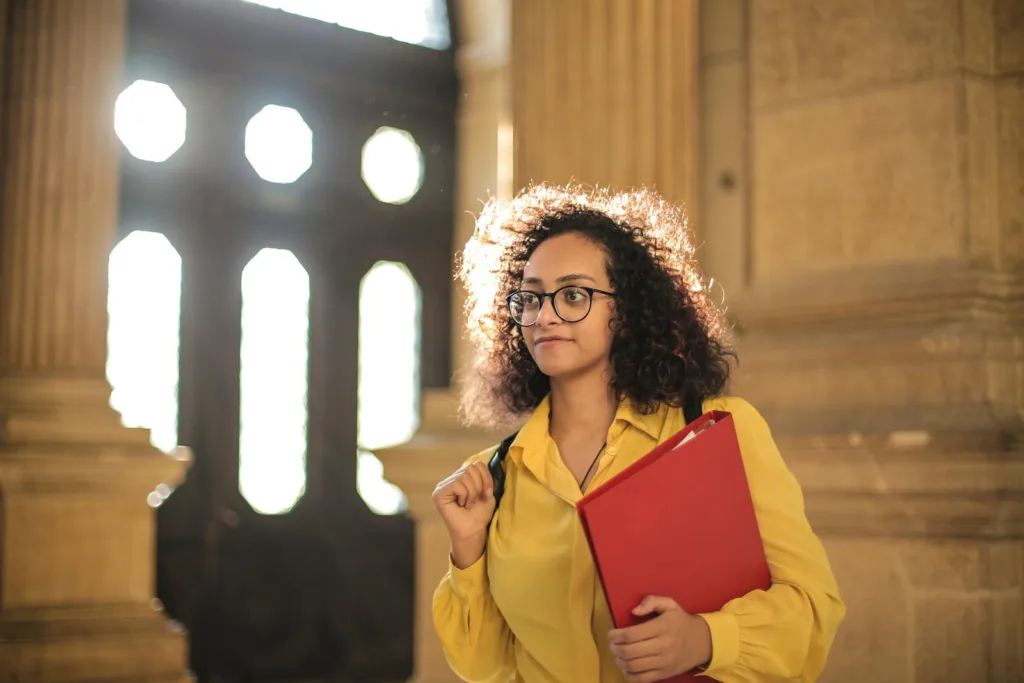 A young woman in a library carrying a red binder, bathed in sunlight.