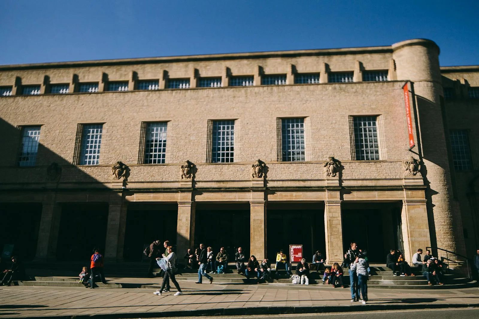 Students relax outside a historic university building under a clear blue sky.