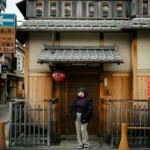 Person standing beside a traditional wooden Japanese building on a street. Captured during the day.